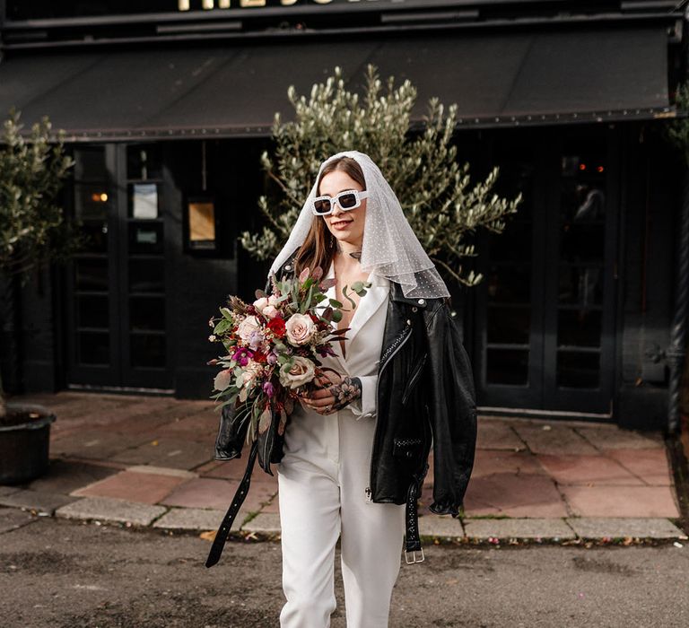 Bride in white jumpsuit with black leather jacket with sparkly white sunglasses and polka dot shoulder length veil 