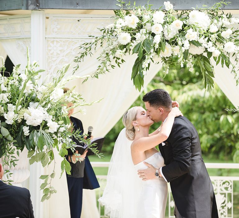 Pretty white and green floral and foliage altar decoration for outdoor wedding ceremony with the bride and groom sharing their first kiss 