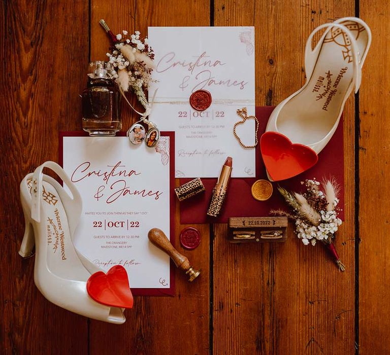 Red and white minimalistic wedding stationery with red cursive calligraphy on white paper and red wax seal with heart shaped Vivienne Westwood wedding shoes, sprigs of dried flowers and foliage, perfume bottles and makeup 