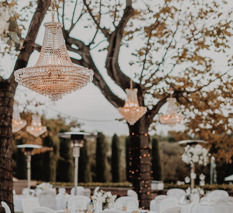 Outdoor wedding classic tablescape  - white tablecloth, bejewelled outdoor chandeliers, fairy lights woven around the trees, peony and foliage table runners, and gold cutlery and crockery 