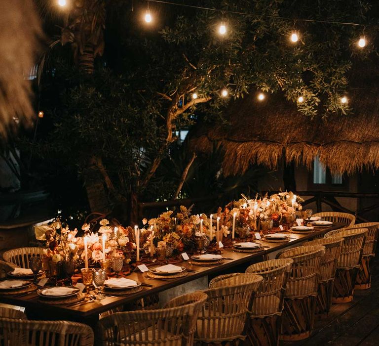 Rustic wedding tablescape by the Mexican sea with wicker chairs, burnt orange dried roses, dried flower and foliage floral centrepieces, wooden slate wedding table numbers, light orange tapered candles and burnt orange table runners