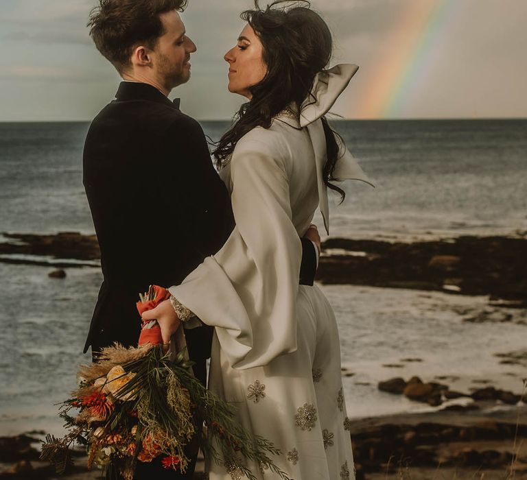 Couple's portrait with rainbow in the background with groom in velvet black suit with bride in sparkly white jumpsuit 