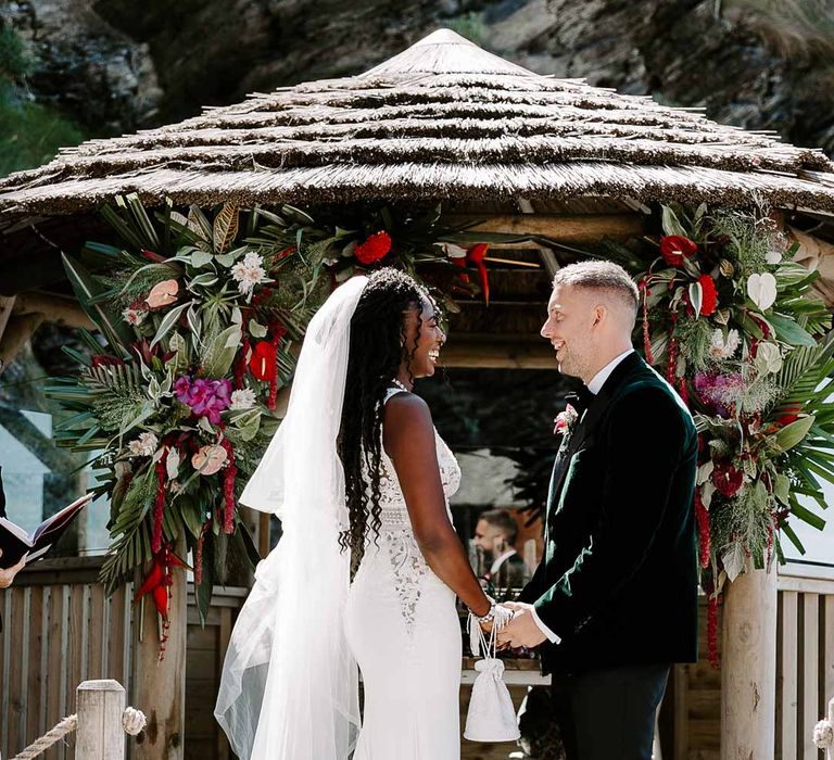 Bride in v neck sleeveless lace Pronovias wedding dress, cathedral length veil and silver sparkly bridal jewellery standing with groom in velvet grooms blazer and black suit trousers 