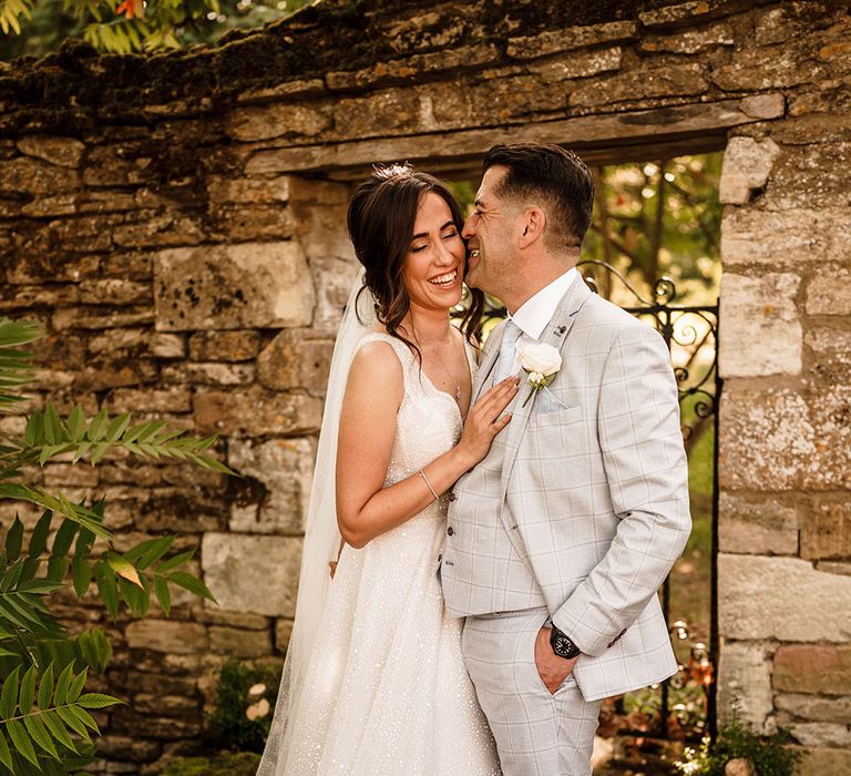 Groom in a light blue check suit kissing his bride in a sparkly wedding dress in the gardens at Caswell House Oxfordshire wedding venue 