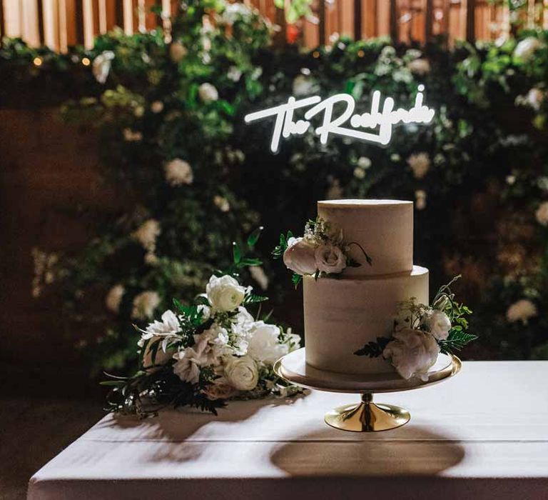 Two tiered buttercream wedding cake with garden rose and foliage decorations on golden cake stand in front of foliage wall and large neon wedding sign