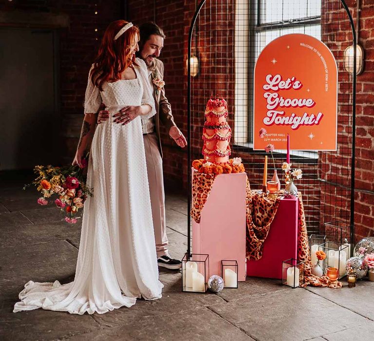 Bride in three quarter length puff sheer sleeve wedding dress with delicate polka dot overlay and square neck and white 3D applique polka dot headband holding large colourful mixed bridal bouquet standing with groom in brown blazer, white shirt, grey suit trousers and vans shoes next to an array of retro wedding decor and signage 