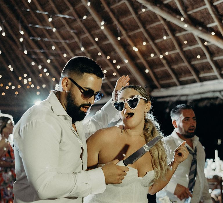 bride and groom in sunglasses cutting their two tier white wedding cake at blue Venado beach wedding reception 