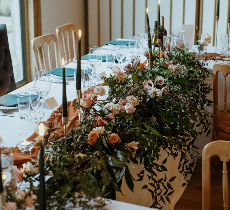 Classic wedding tablescape with white tablecloth, burnt orange wedding tablerunner, forest green wedding napkins, forest green tapered candles and bottles of wine with large light pink and orange garden rose and foliage table centrepiece