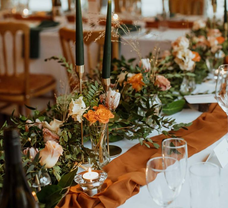 Burnt orange wedding tablerunner, forest green wedding napkins, forest green tapered candles and bottles of wine with suspended foliage and fairy lights decor at wedding