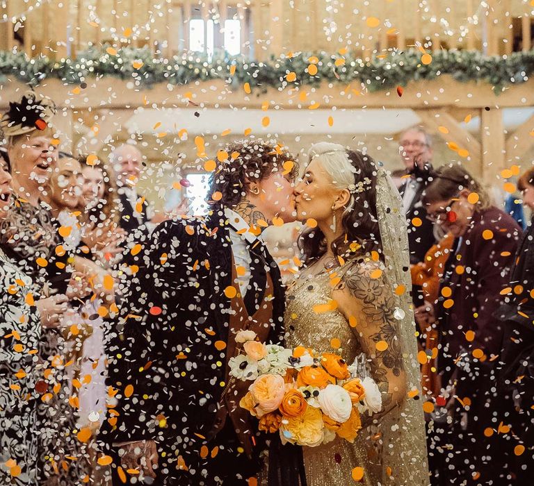 Bride in dark velvet bridal jacket with rust waistcoat and pocket square, black suit trousers and gold pocket watch kissing bride in sleeveless deep v-neck sparkly gold wedding dress with sheer and gold polka dot wedding veil holding orange garden rose, white poppy, light pink carnation and foliage bridal bouquet wrapped in sparkly gold ribbon during colourful confetti exit at Kingscote Barn Tetbury wedding venue with fairy lights and festoon lighting