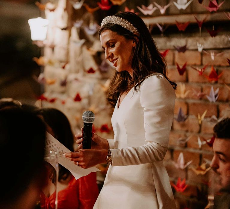 Bride in long sleeve short wedding dress and pearl bridal headband doing bridal speech standing in front of origami paper cranes DIY wedding decor at Willow Marsh Farm Loughborough 