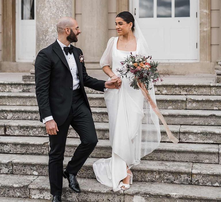 Groom in classic black tuxedo with bowtie and wildflower boutonniere walking hand in hand with bride in short sheer puff sleeve wedding dress with square neck and wrist loop with sheer pearl embellished bridal veil holding dried wildflower bouquet at Buxted Park