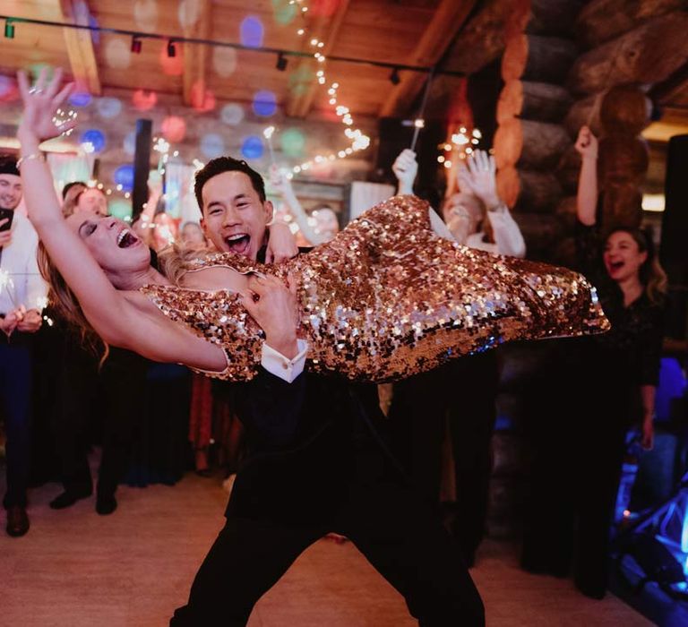 Groom in classic black tuxedo, black bowtie and white garden rose boutonniere picking up bride in gold sequin sparkly reception dress during fun wedding dance at La Cabane Des Praz 