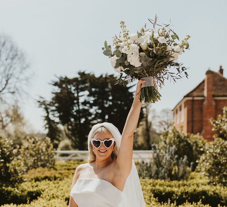 Bride in strapless Eva Lendel wedding dress raising white rose wedding bouquet in the air wearing white heart retro wedding sunglasses 