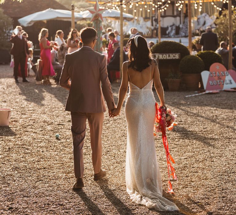 Bride in low back sparkly Made With Love wedding dress and headband walking hand in hand with the groom in a taupe suit 