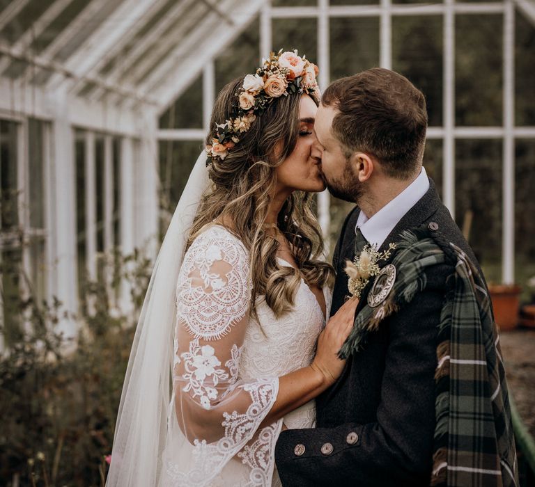 Boho bride in rose flower crown and veil kisses groom in tartan in greenhouse at highland wedding in Glencoe