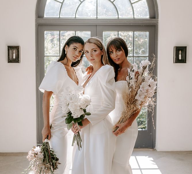 Bridesmaids in different white dresses and jumpsuits holding rose and dried flower bouquets 