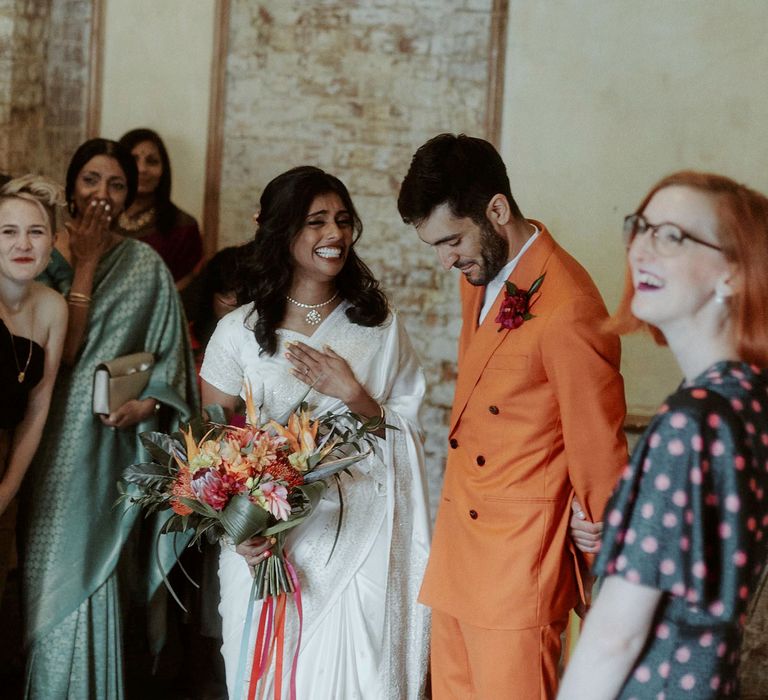 Bride in a white sari holding a tropical flower bouquet laughing with her husband in a double breasted orange wedding suit 