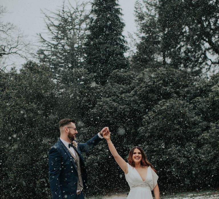 Groom in navy suit and bow tie dances with bride in white dress with silver detailing outside at snowy wedding