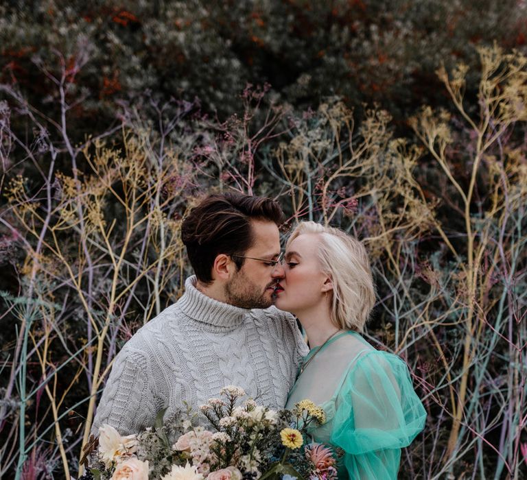 Groom in a grey turtle neck jumper kissing his bride in a pastel green sheer wedding dress on the beach 