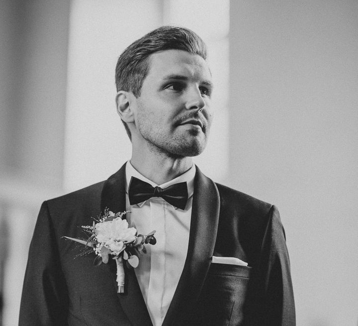 Groom in Tiger of Sweden tuxedo and black silk tie with white buttonhole waits for bride at the altar during German wedding