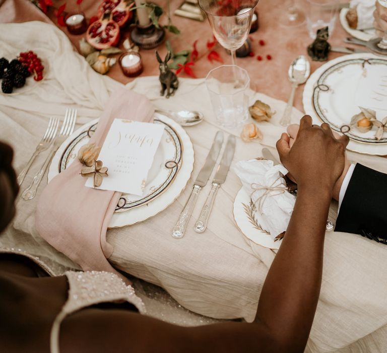 Bride and groom holding hands at their sweetheart table with ornate tableware, silver cutlery and natural linens 