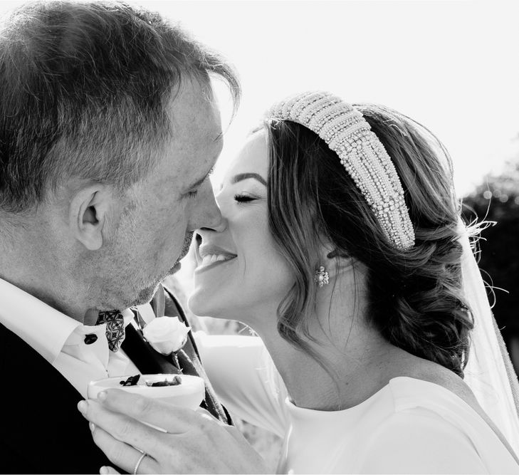 Smiling bride in bridal headband kisses groom in navy suit with white rose buttonhole whilst holding cocktail