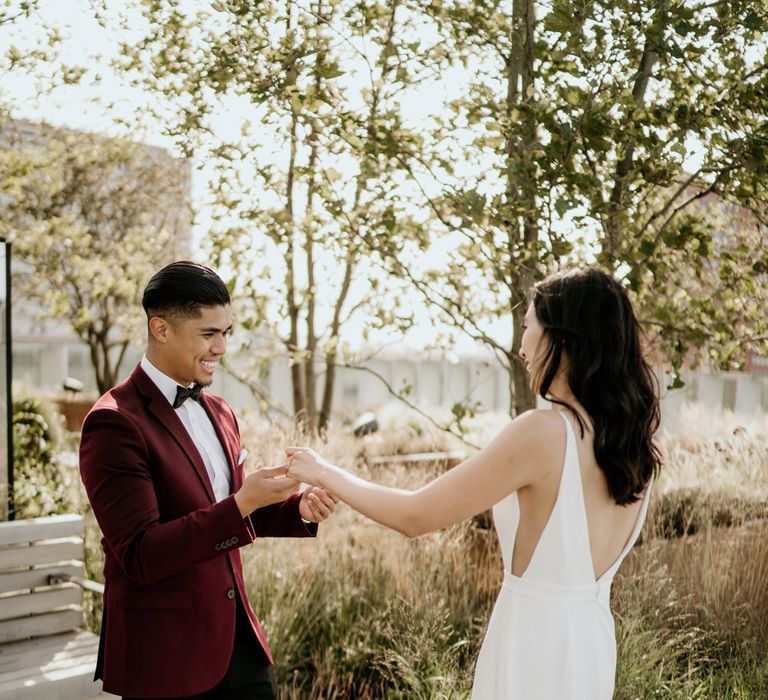 Bride and groom first look with groom in a burgundy tuxedo jacket and bow tie and bride in a fitted Sash Seven wedding dress with sheer side panels 