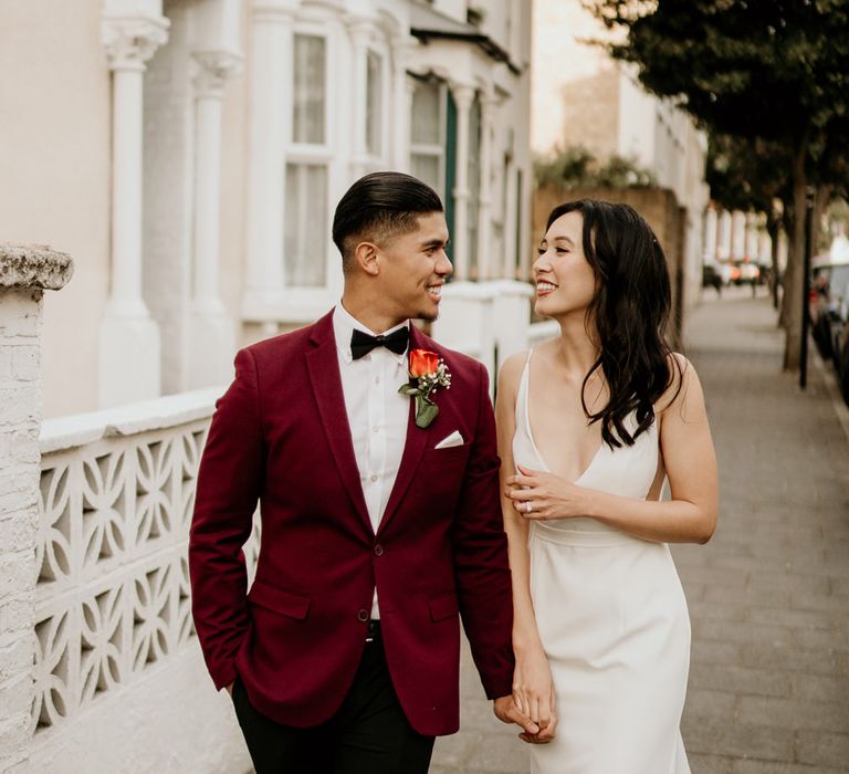 Filipino bride and groom in a minimalist wedding dress and dark red tuxedo jacket walking through London 