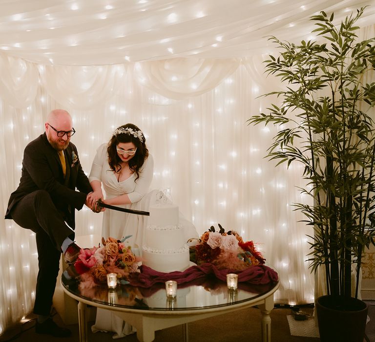 Bride in white Rime Arodaky wedding dress with bridal headband and white cat eye glasses and groom in navy suit and yellow tie cut cake with sword