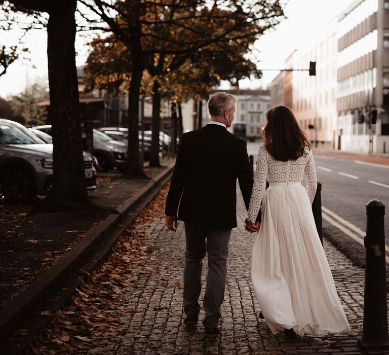 Bride in long sleeved lace top Self Portrait wedding dress holds hands with groom in brown woollen blazer walking through cobbled streets of Bristol