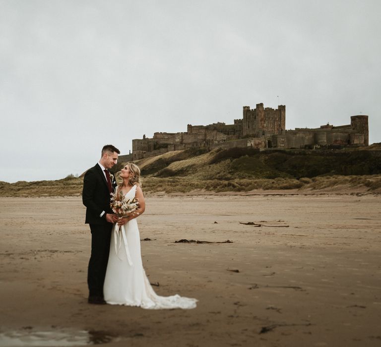 Bride & groom stand together on the beach with Bamburgh Castle in the background