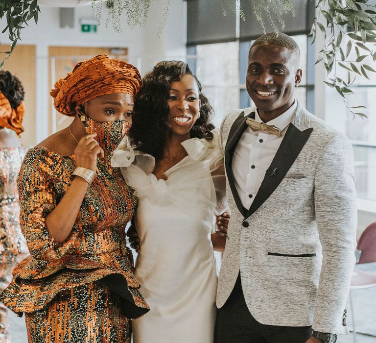 Bride in white satin wedding dress with shoulder bows stands in between wedding guests in orange and black printed dress, headscarf and face mask and groom in grey suit jacket with black lapels at Bridge Community Church wedding