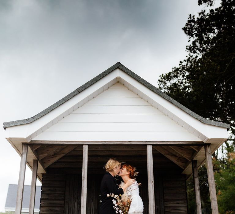 Two brides kiss under beach hut on overcast day