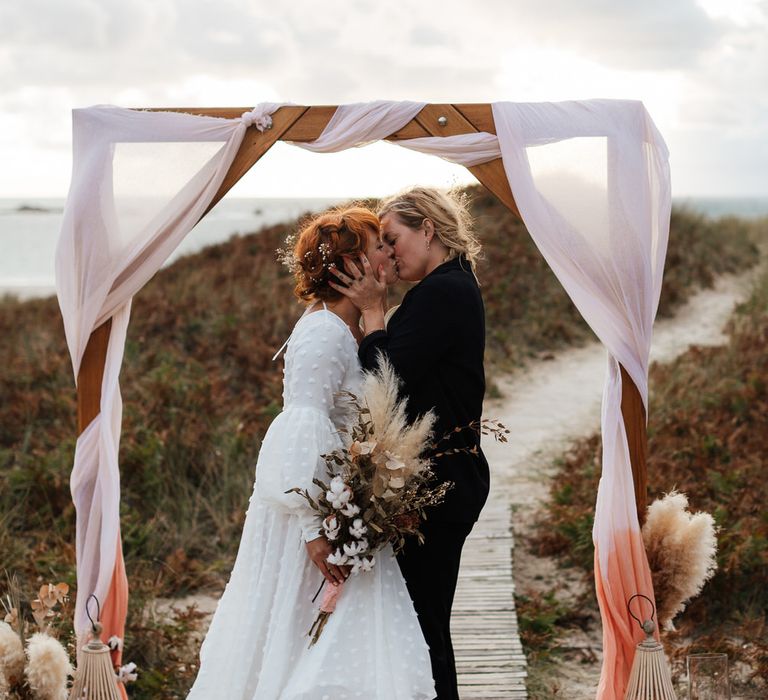 Brides kiss under wooden arch at beach boho elopement