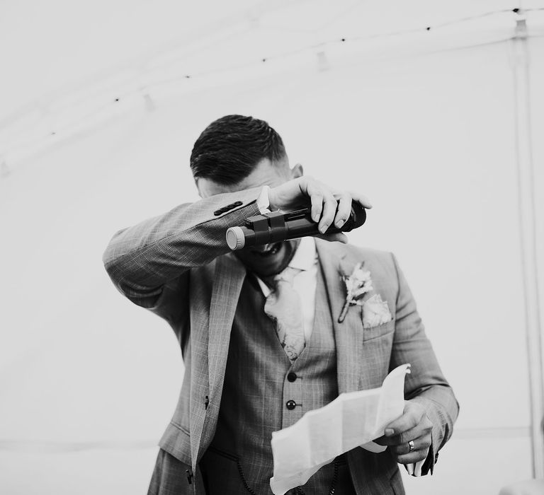 Groom in grey Hugo Boss suit with waistcoat and yellow tie holds arm over face as he reads speech