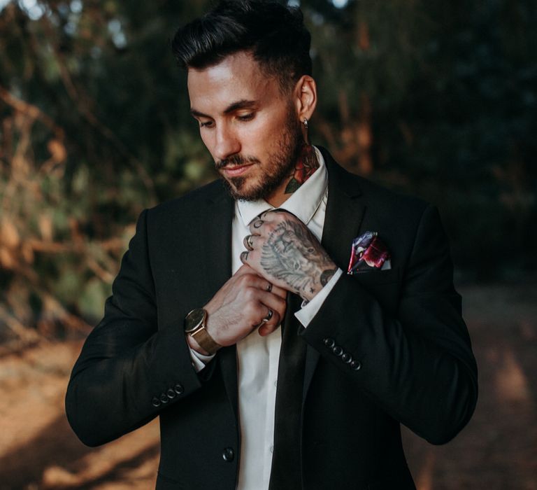 Groom with tattoos fixing his tie in the woods