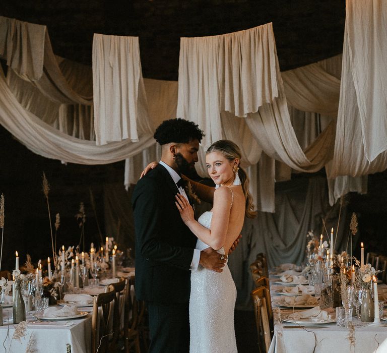 Groom in a tuxedo and bride in a sequin wedding dress standing in their Pentney Abbey wedding reception with ceiling drapes 