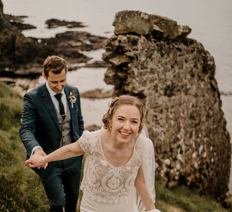 Smiling bride in lace top capped sleeve wedding dress with satin skirt holds hands of groom whilst they walk up hill after Dunluce Castle wedding