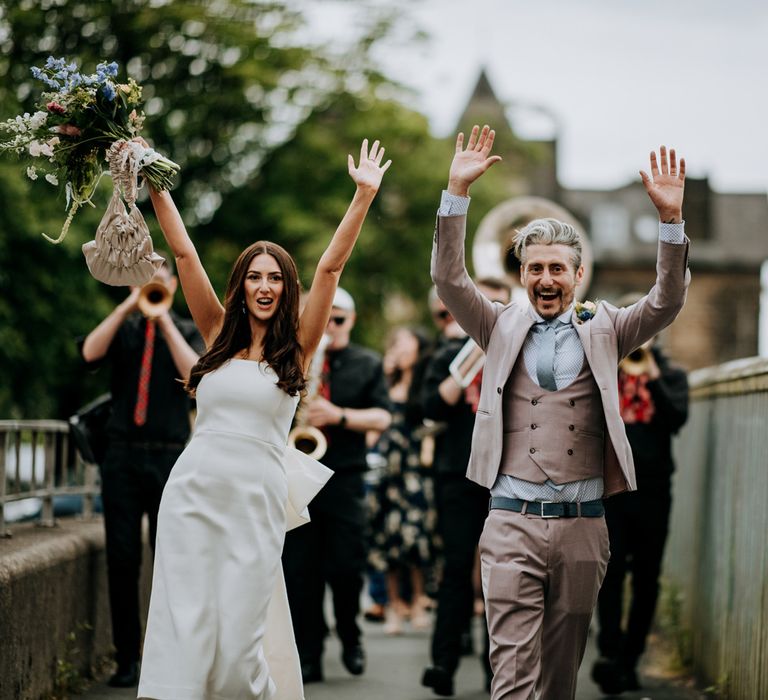 Bride in white strapless Rebecca Vallance Dress and groom in brown Moss Bros suit with blue tie throw hands in the air as they're followed by the New York Brass Band around Harrogate after wedding ceremony