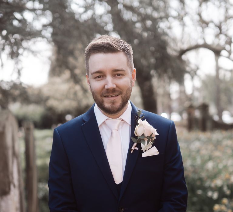 Smiling groom in a navy suit with pastel pink tie, and rose lapel flower