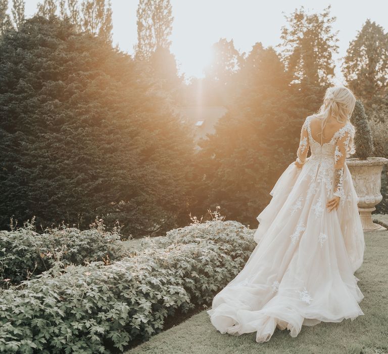 A blonde bride walks away from the camera in long tulle layered wedding dress as the sun sets.