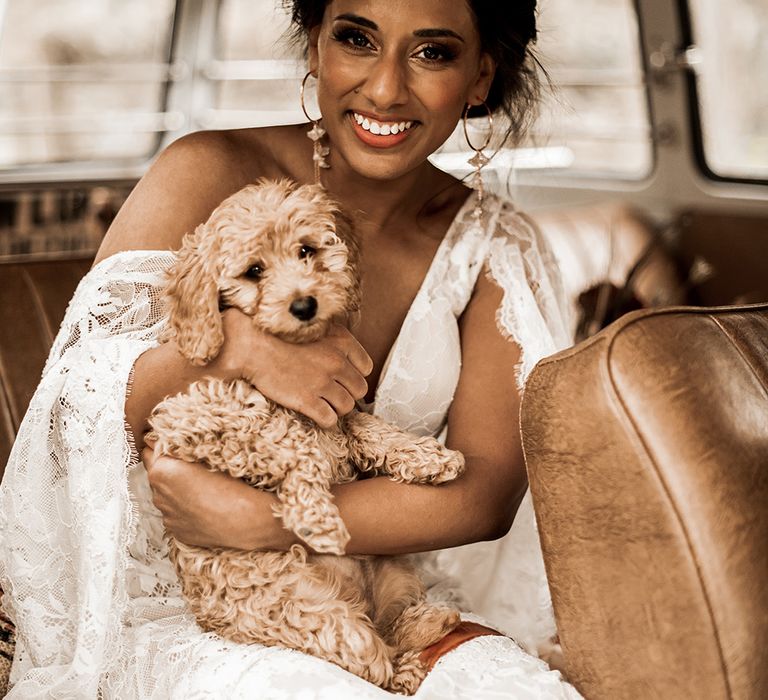 Indian bride in a lace wedding dress and hoop earrings holding a tan cockapoo puppy 