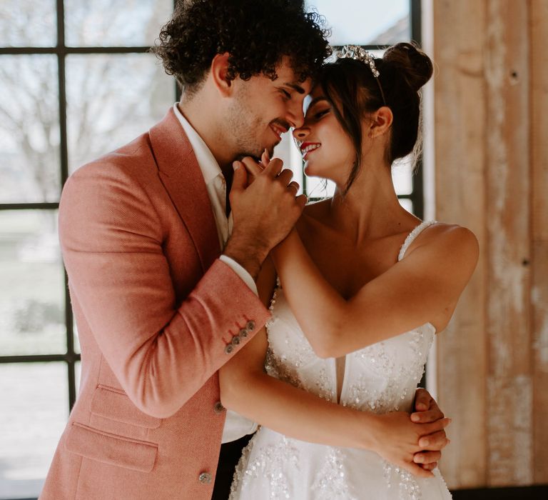 Bride in an embellished wedding dress with full skirt, fitted bodice and thin straps being embraced by her groom in a red tweed jacket 