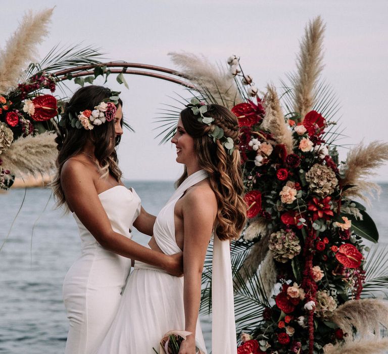 Two brides on the beach in front of altar flowers of red anthuriums, roses, pampas grass, dried hydrangea, pink carnations, eucalyptus and palm leaves