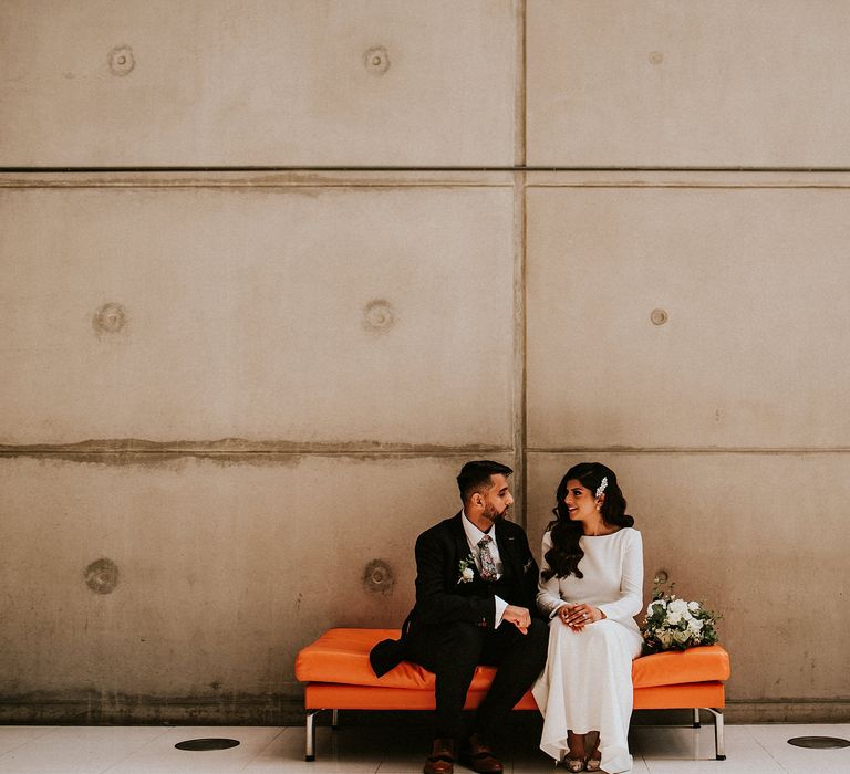 Bride in long sleeve wedding dress sitting on an orange bench with her husband at Brent Civic Centre wedding venue