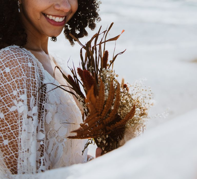 Bride looks at camera smiling broadly as she walks along the beach front whilst holding bouquet of dried flowers