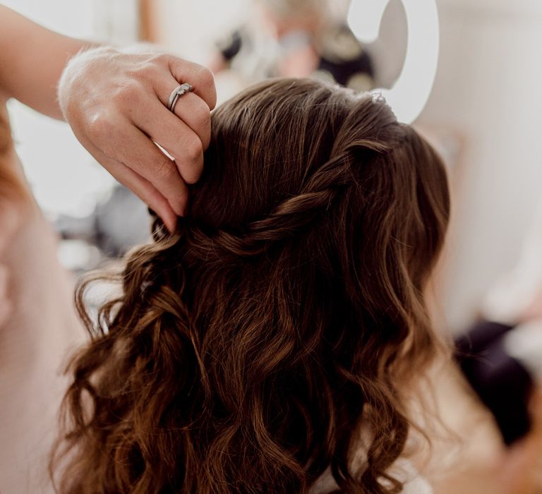 Bride with long brown curled hair has braid pinned up by hairdresser before wedding