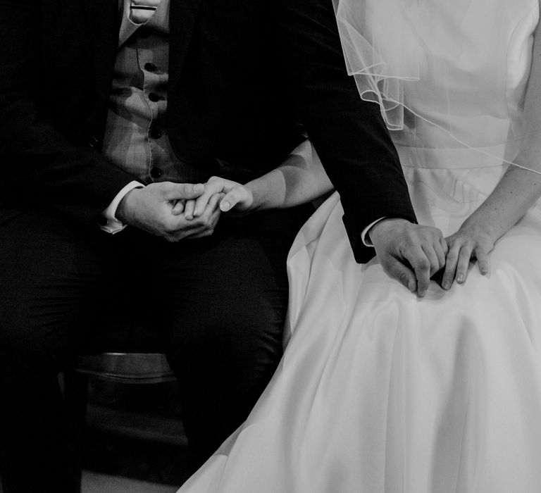 Groom in three piece suit and bride in white Elbeth Gillis gown and veil sit holding hands in church during wedding ceremony