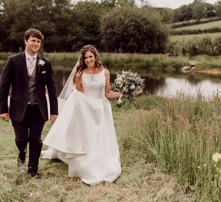 Bride in white Elbeth Gillis wedding dress and veil holding white and green bridal bouquet walks through field with groom in dark brown three piece suit with grey waistcoat at home farm wedding